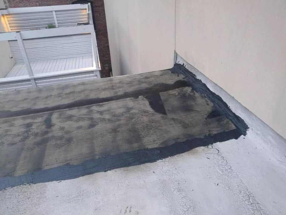 Flat rooftop corner with weathered waterproofing and a window beside a beige wall.