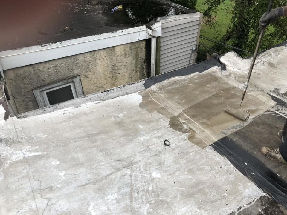 Flat rooftop with white coating, chimney, and nearby building wall beside trees.