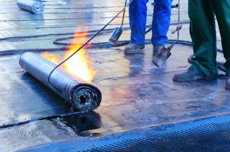 Roofing torch melting a rolled membrane on a flat roof with workers nearby