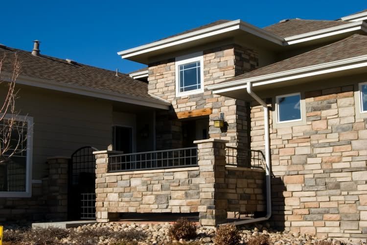 Stone house exterior with front porch, metal railing, and brown shingle roof in daylight