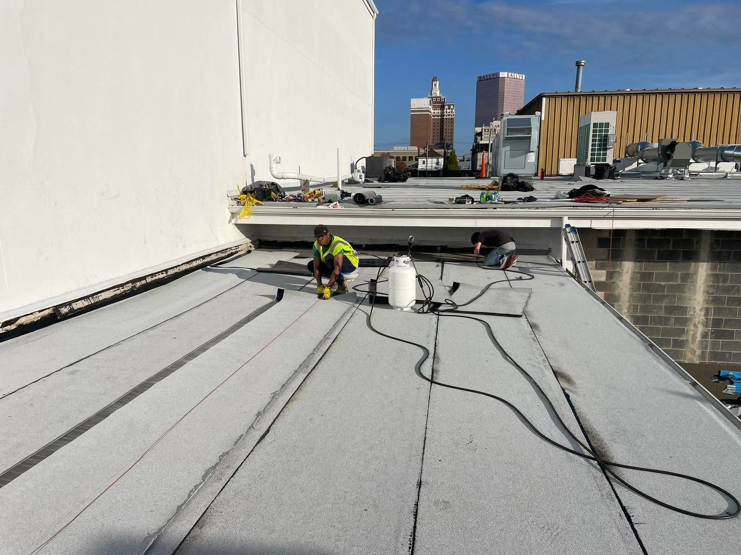 Workers on a flat rooftop installing or repairing roofing membrane near city buildings.