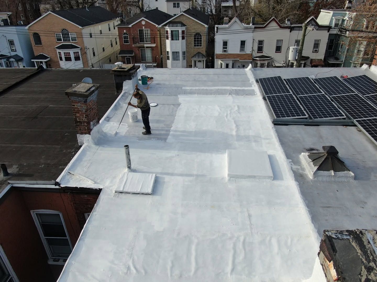 Worker on a white rooftop applying coating near solar panels, with houses in the background