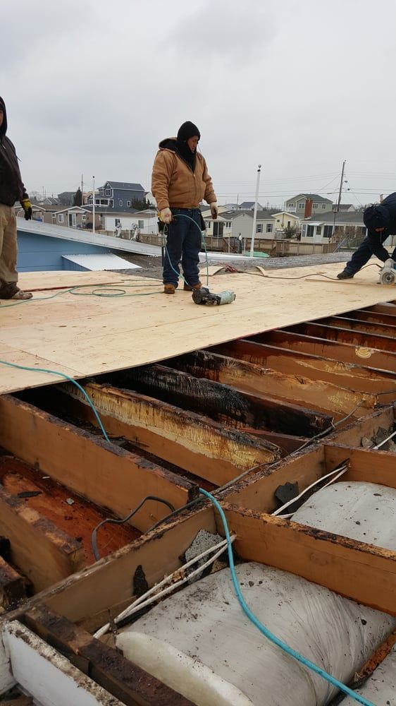 Workers installing plywood sheathing on a rooftop over exposed beams and wiring under a cloudy sky