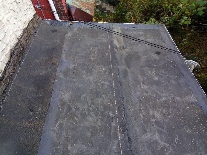 Flat weathered rooftop with seams beside a brick wall and greenery beyond