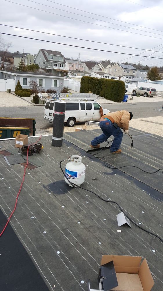 Worker kneeling on a rooftop, installing black roofing underlayment with tools and buckets nearby.