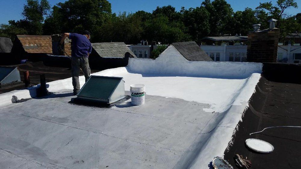 Worker applying white roof coating on a flat rooftop with buckets and tools.