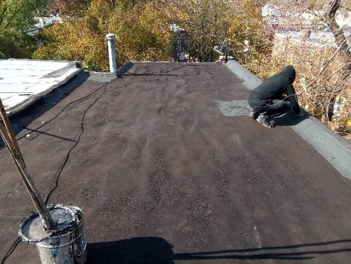 Worker applying roofing material on a flat roof near trees and a chimney