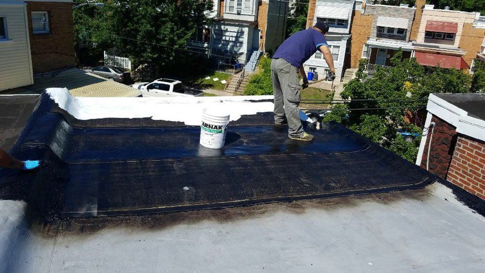 Worker applying black roofing material on a flat roof near a white bucket