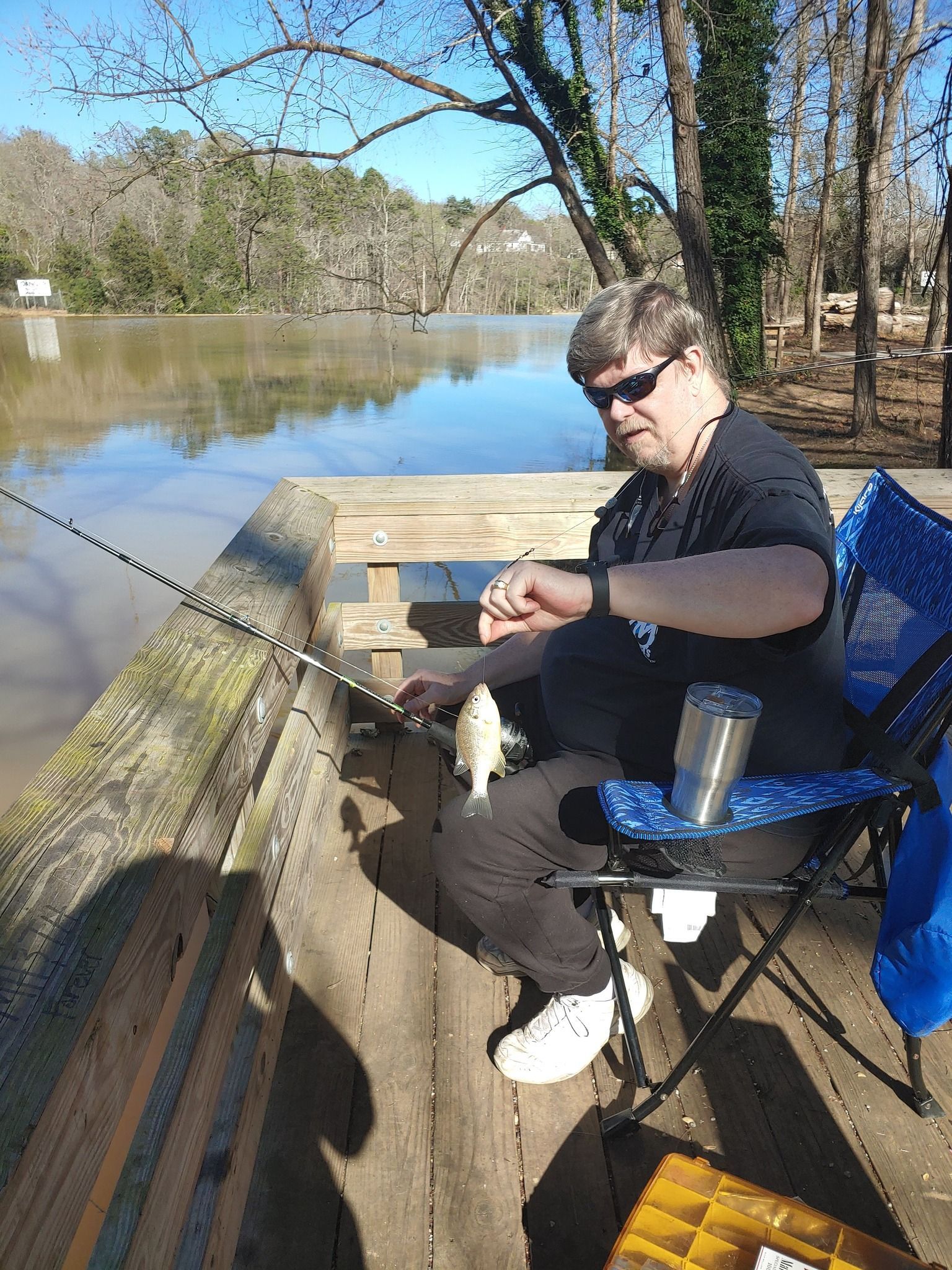 Man fishing from a wooden dock, holding a caught fish. He is wearing sunglasses, and sitting in a chair. Water in the background.