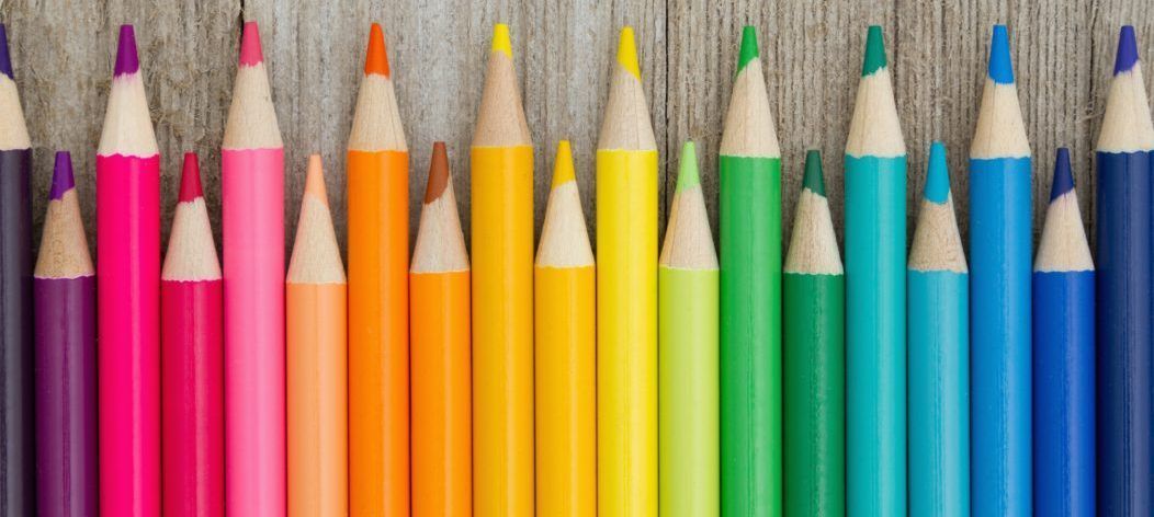 Row of colored pencils arranged in a rainbow spectrum against a wooden background.