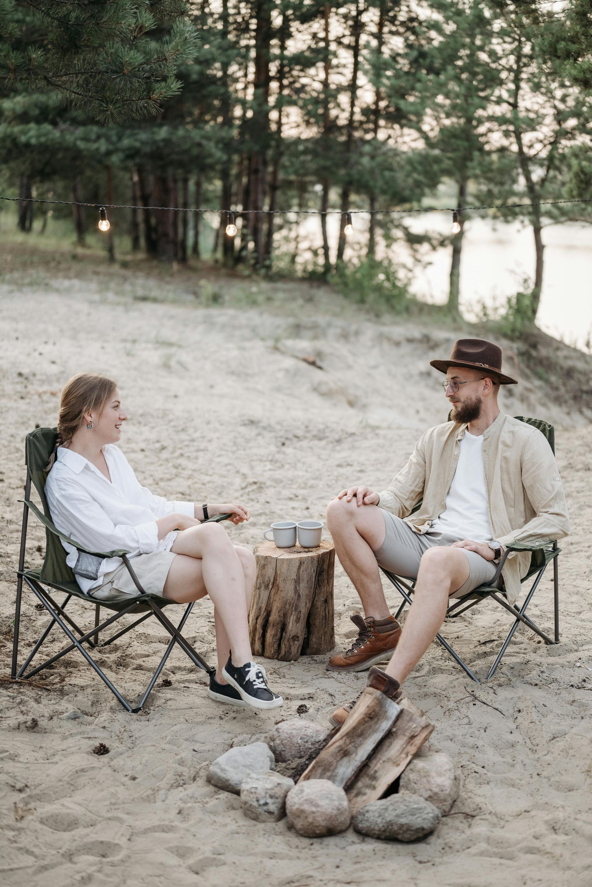 Couple sitting in camping chairs by a fire on a sandy beach, with trees in the background.