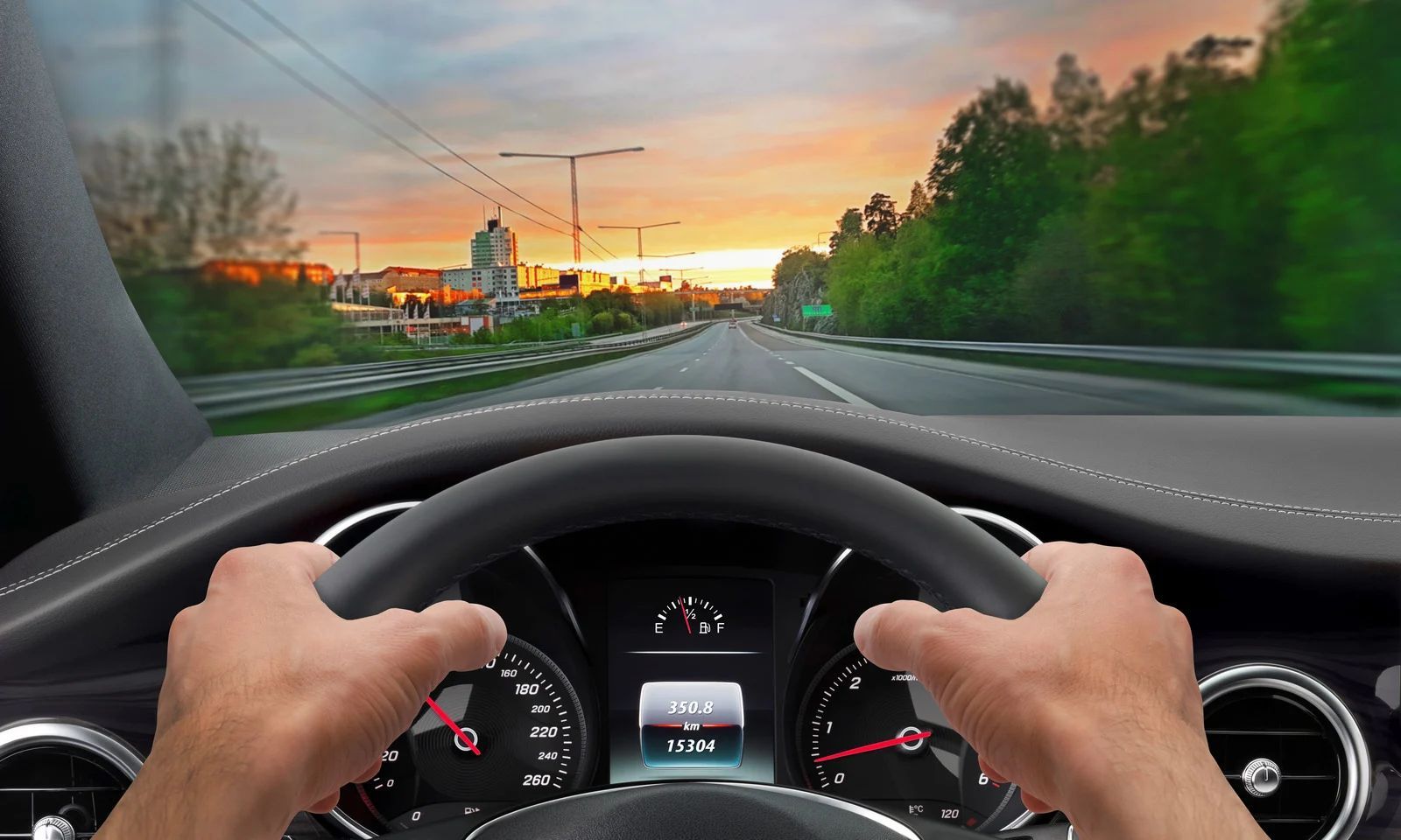 Driver’s view of hands on a steering wheel, driving on a highway at sunset with trees and road ahead