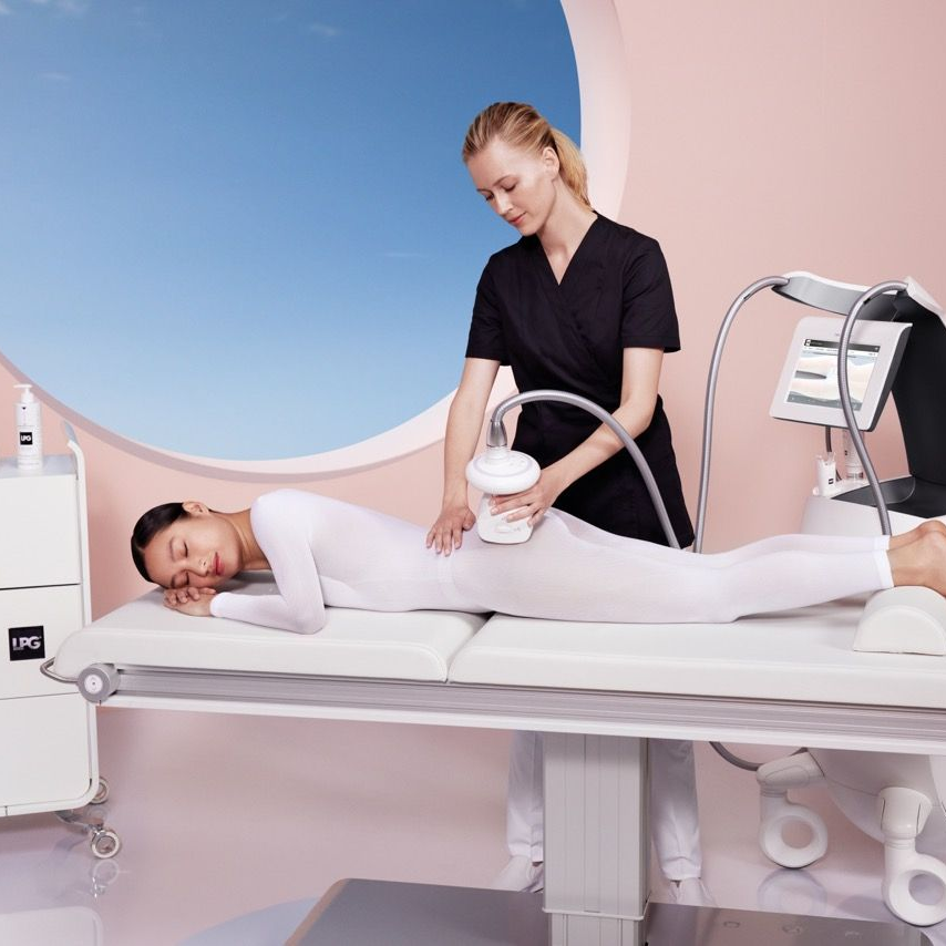 Femme recevant un soin corporel sur une table de spa ; technicienne utilisant l'appareil. Chambre rose clair, vue sur le ciel bleu.