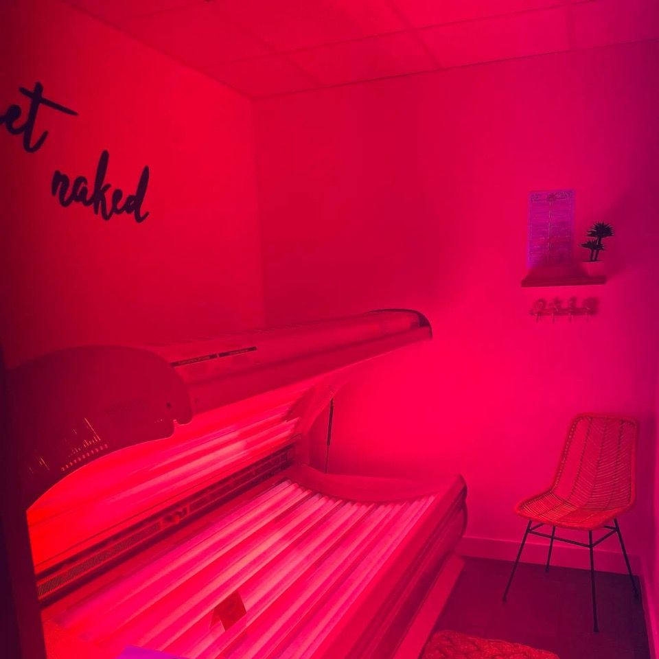 Un solarium dans une pièce éclairée en rose, avec un panneau « Déshabillez-vous ». Une chaise et une étagère avec une plante complètent le décor.