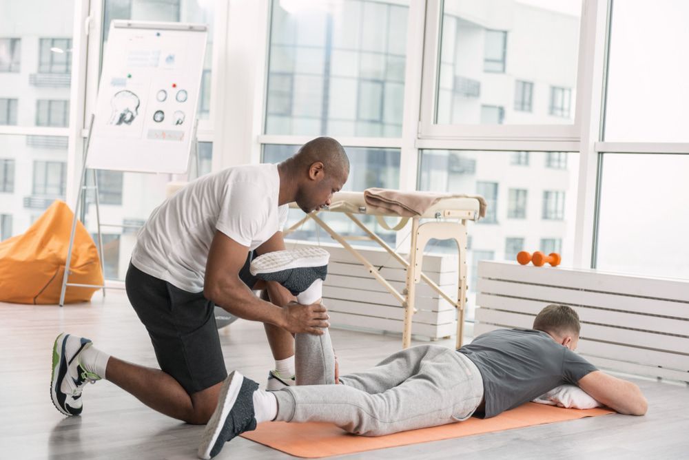 A Man Is Helping Another Man Stretch His Leg On A Yoga Mat — Physiocare In Buderim, QLD