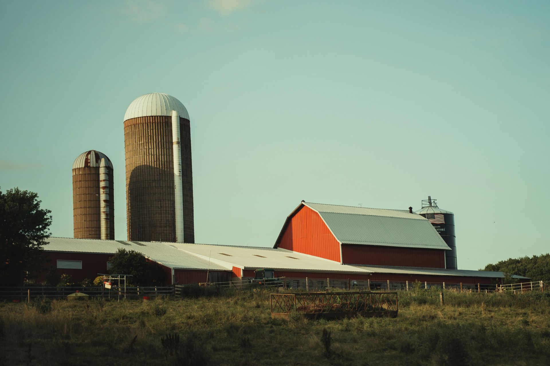 A red barn with two silos against a pale blue sky, set on a grassy farm field.