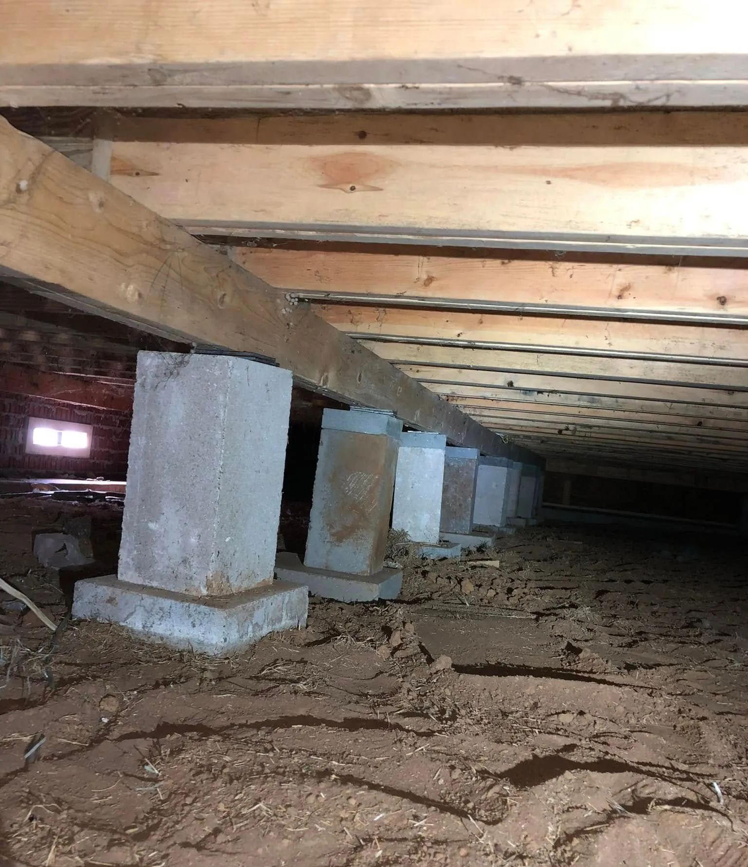 Crawlspace beneath a house with concrete support piers, exposed beams, and dirt floor