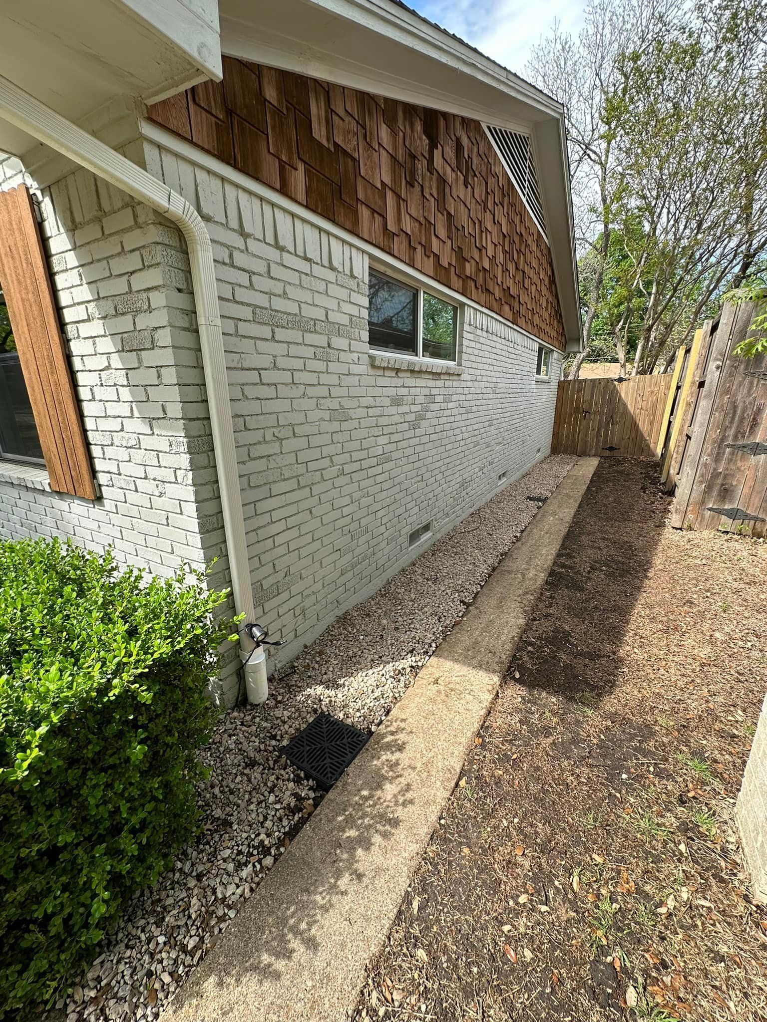 Side yard beside a white brick house with a narrow concrete walkway and mulch under eaves