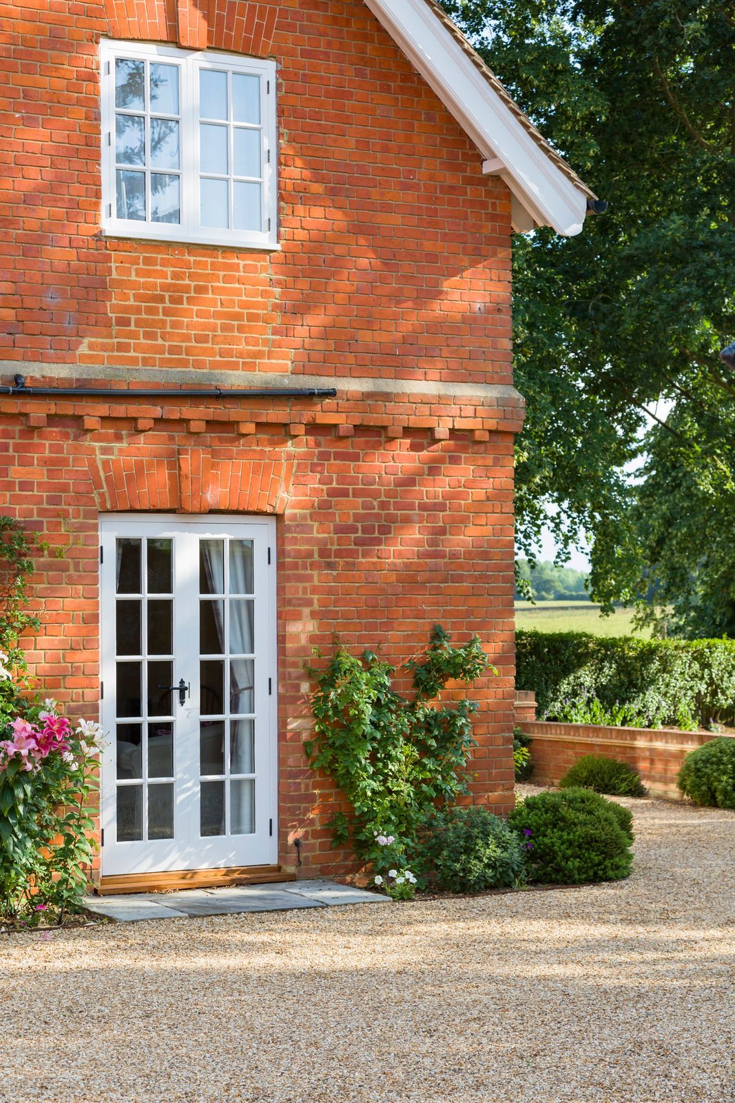 Red brick cottage with white French doors and a small upper window beside a gravel path and garden greenery