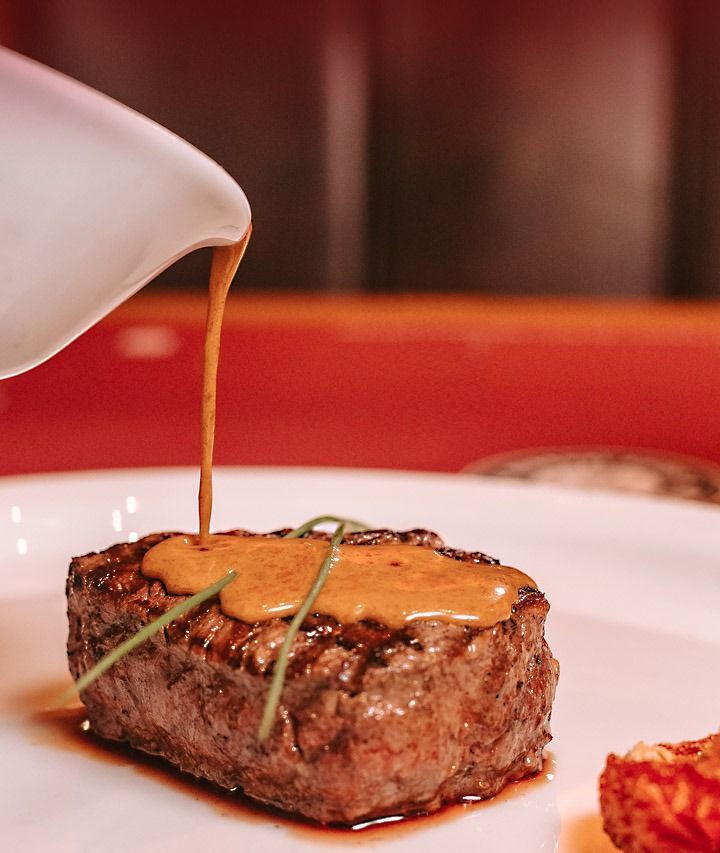 Molho sendo derramado sobre um bife grelhado em um prato branco em um restaurante.