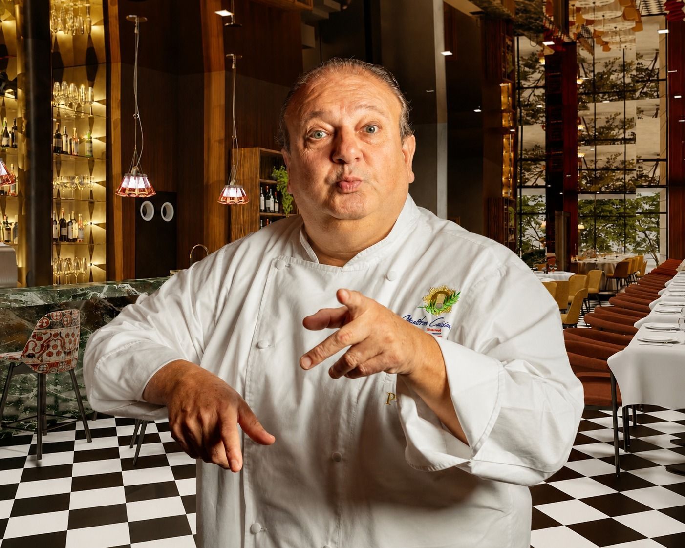 Chef de cozinha de jaleco branco gesticulando em um restaurante com piso quadriculado em preto e branco.