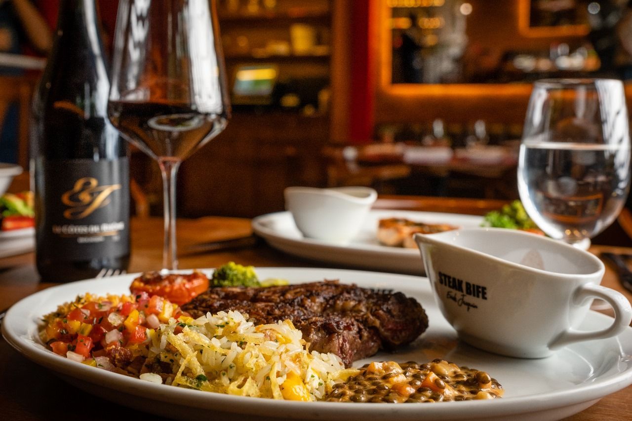Jantar com bife servido à mesa, acompanhamentos e taças de vinho em um restaurante.
