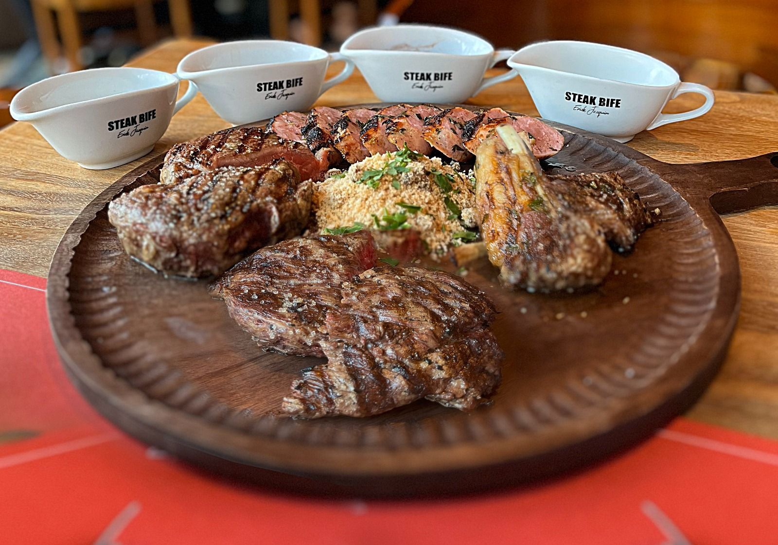 Bifes grelhados e acompanhamentos em uma travessa de madeira com pequenas xícaras em um ambiente de restaurante.