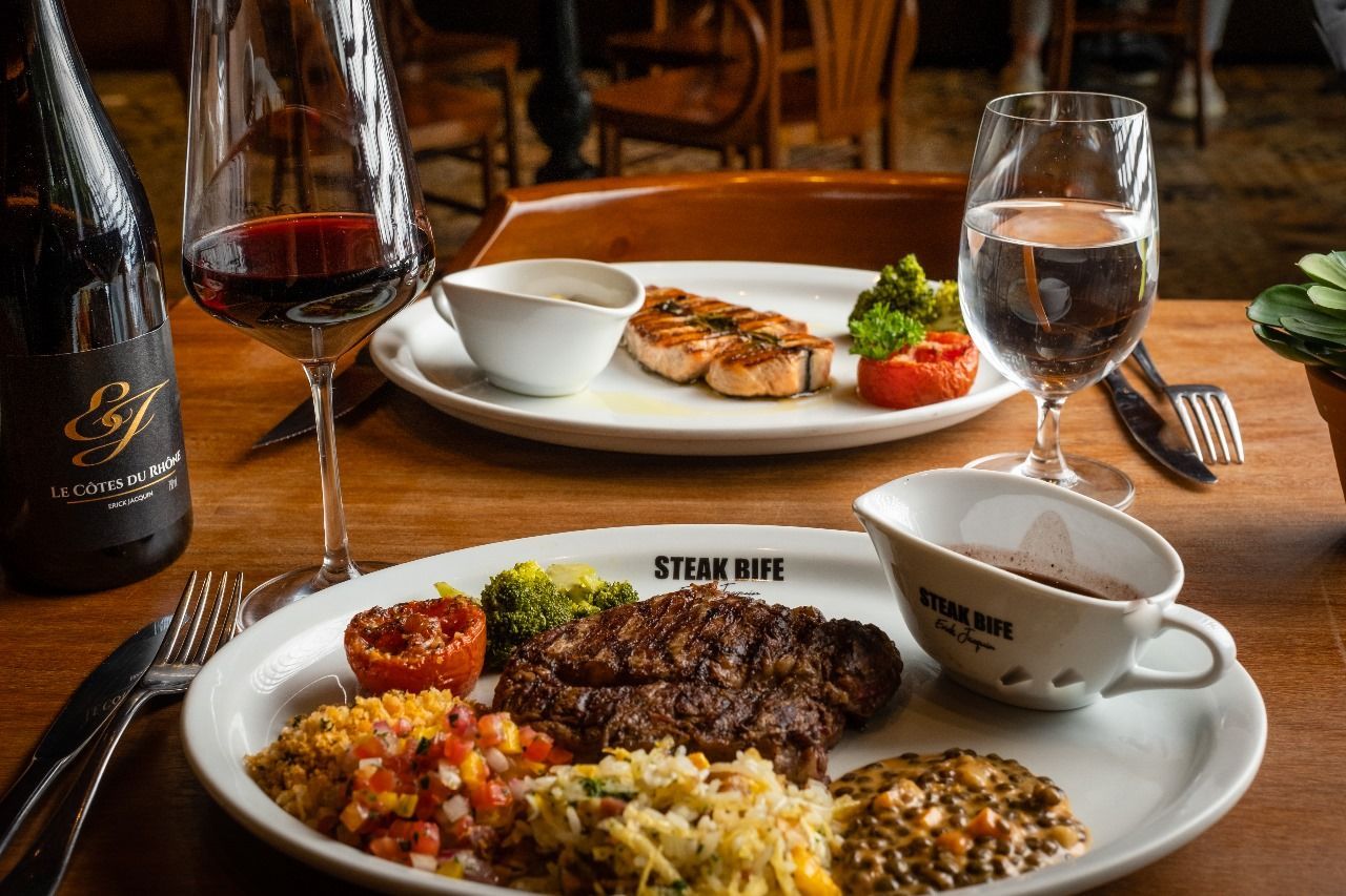 Mesa de restaurante com bife e acompanhamentos, taças de vinho e um aperitivo servido ao fundo.