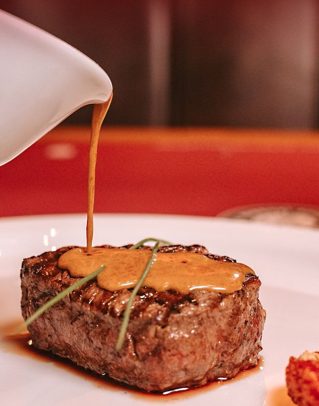 Bife em um prato branco sendo coberto com molho marrom retirado de uma jarra.