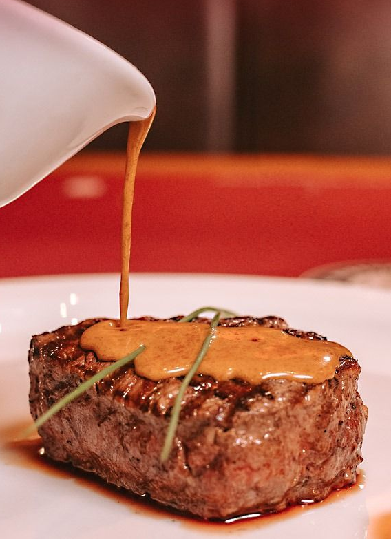 Bife servido em um prato branco, coberto com molho marrom retirado de uma jarra branca.