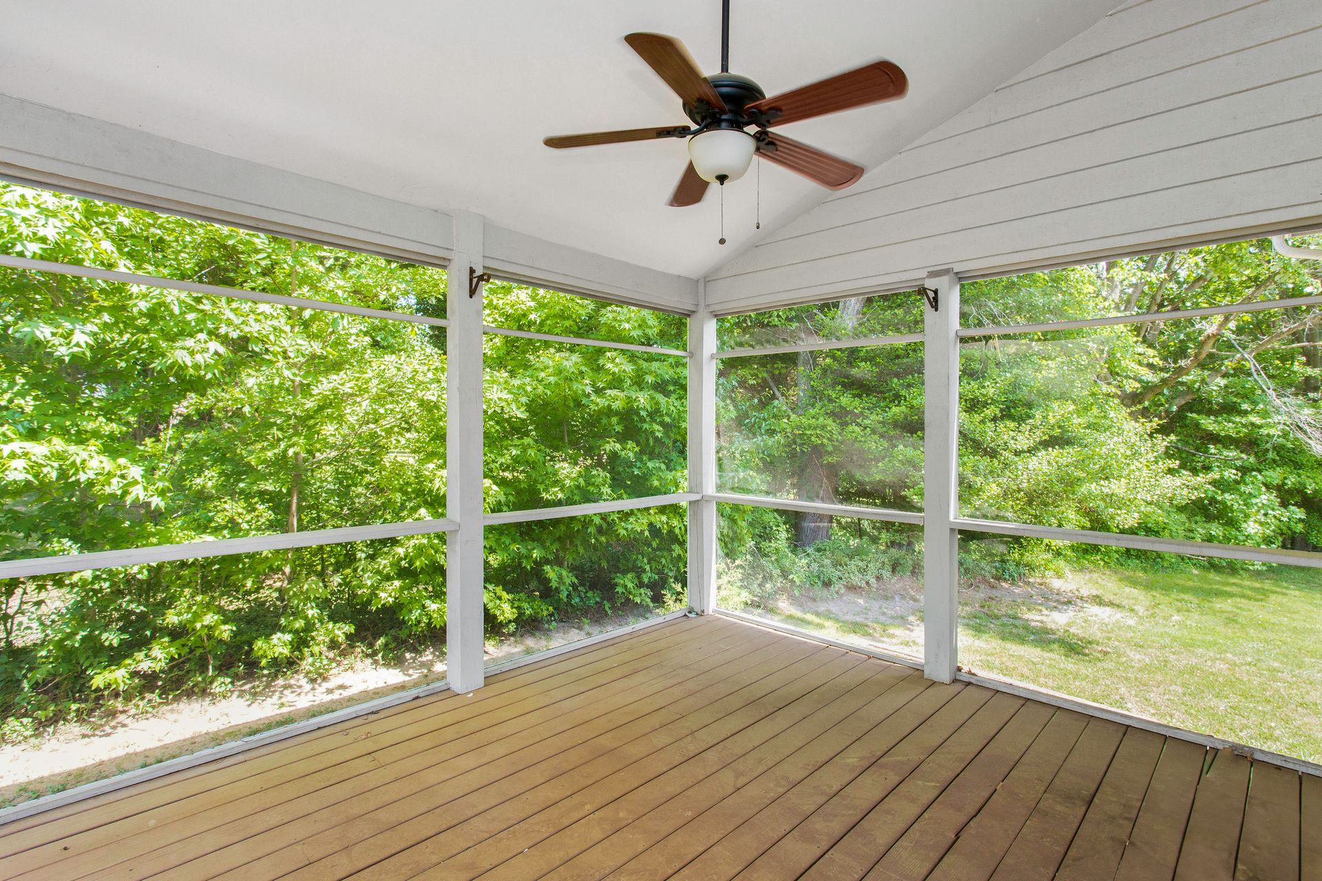 Screened-in porch with a wooden floor, white pillars, and a ceiling fan; surrounded by lush green trees.