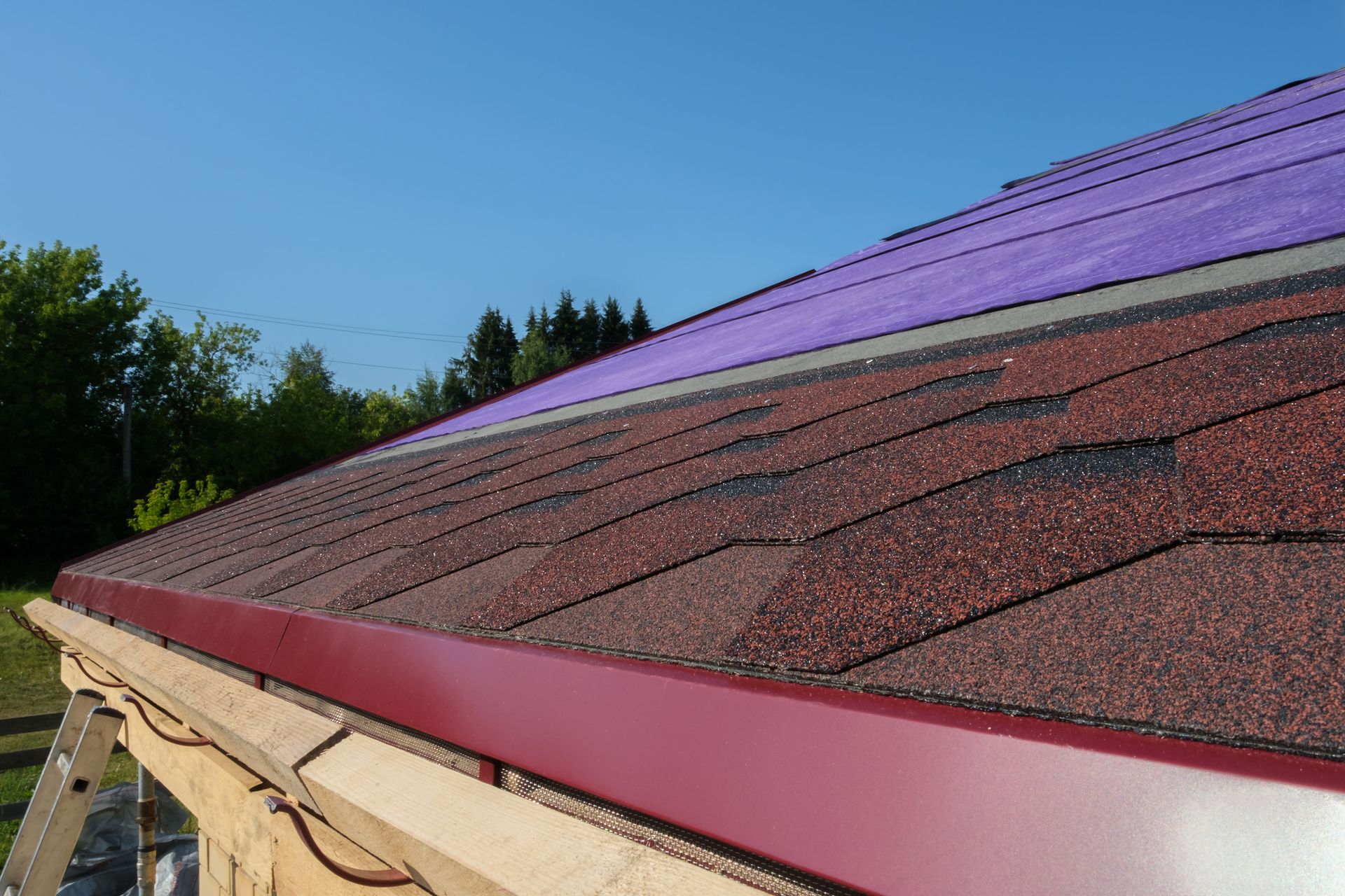 Close-up of a roof with red shingles, a burgundy gutter, and purple underlayment. The setting is outdoors on a sunny day.