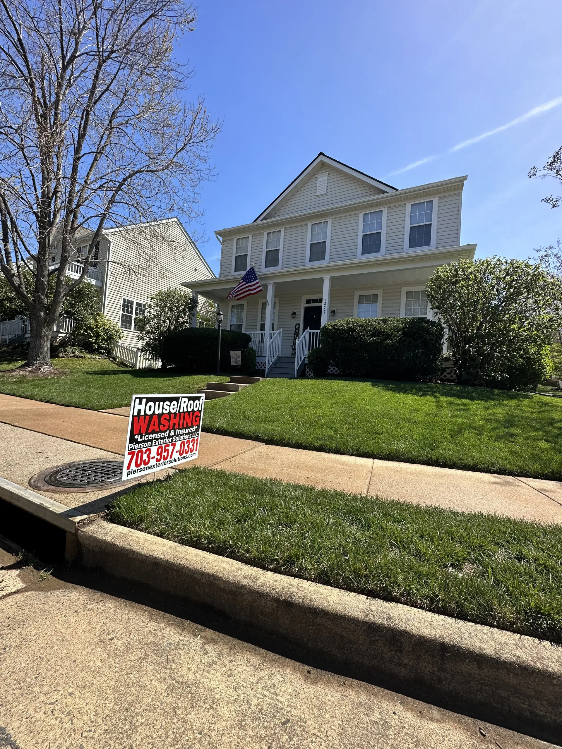 Front yard with a yellow house, bare tree, and a political yard sign by the sidewalk under a clear blue sky.
