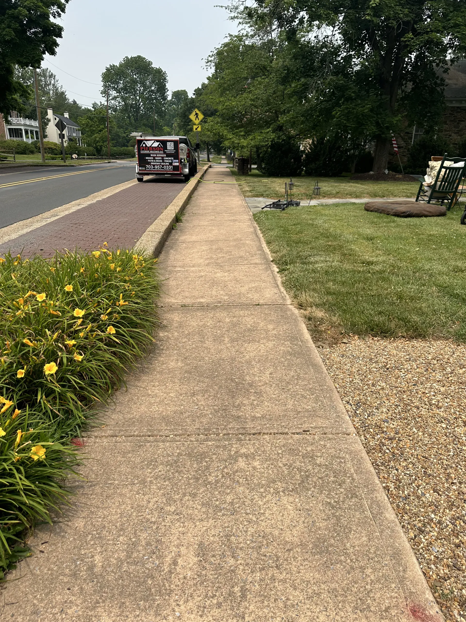 Sidewalk beside a road with a parked black vehicle, trees, grass, and yellow flowers under a cloudy sky