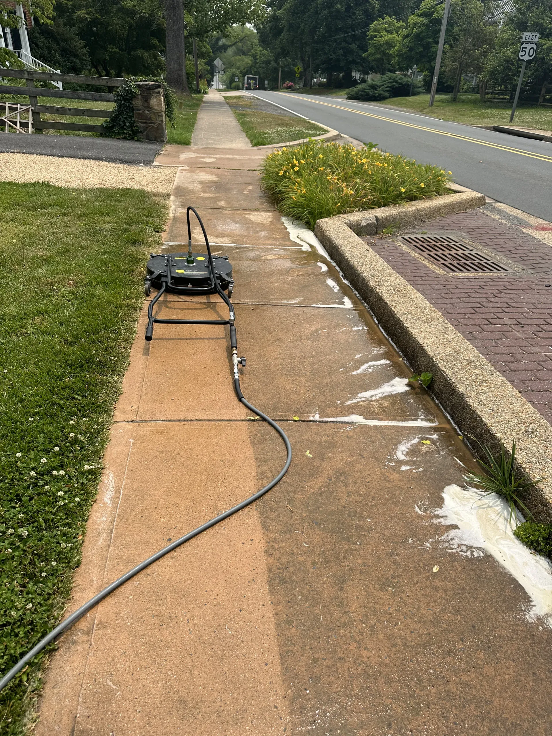 A black device on a sidewalk with a long cable, beside a road and grass in a suburban area