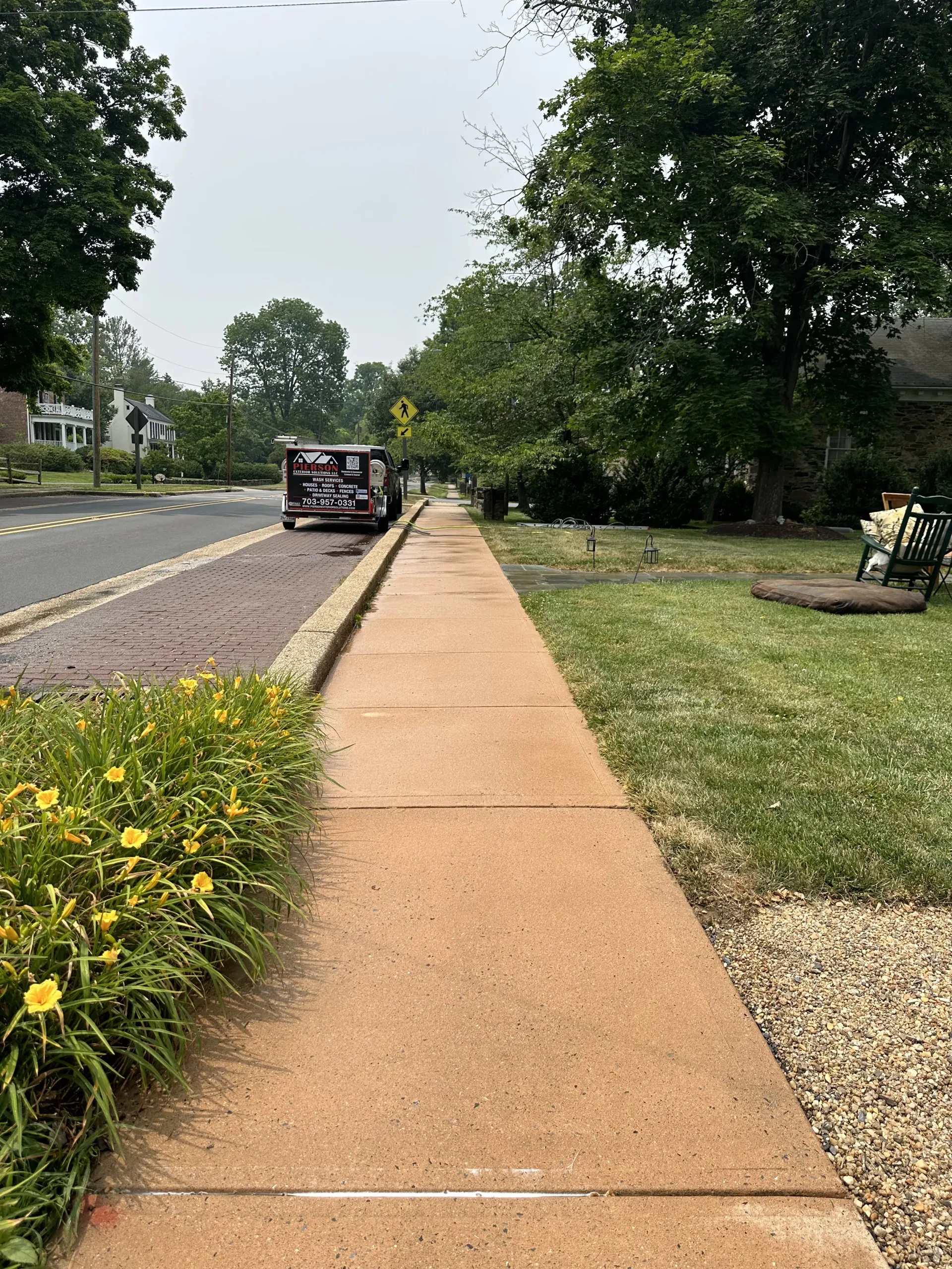 Sidewalk beside a road with a parked vehicle, trees, and yellow flowers along the edge