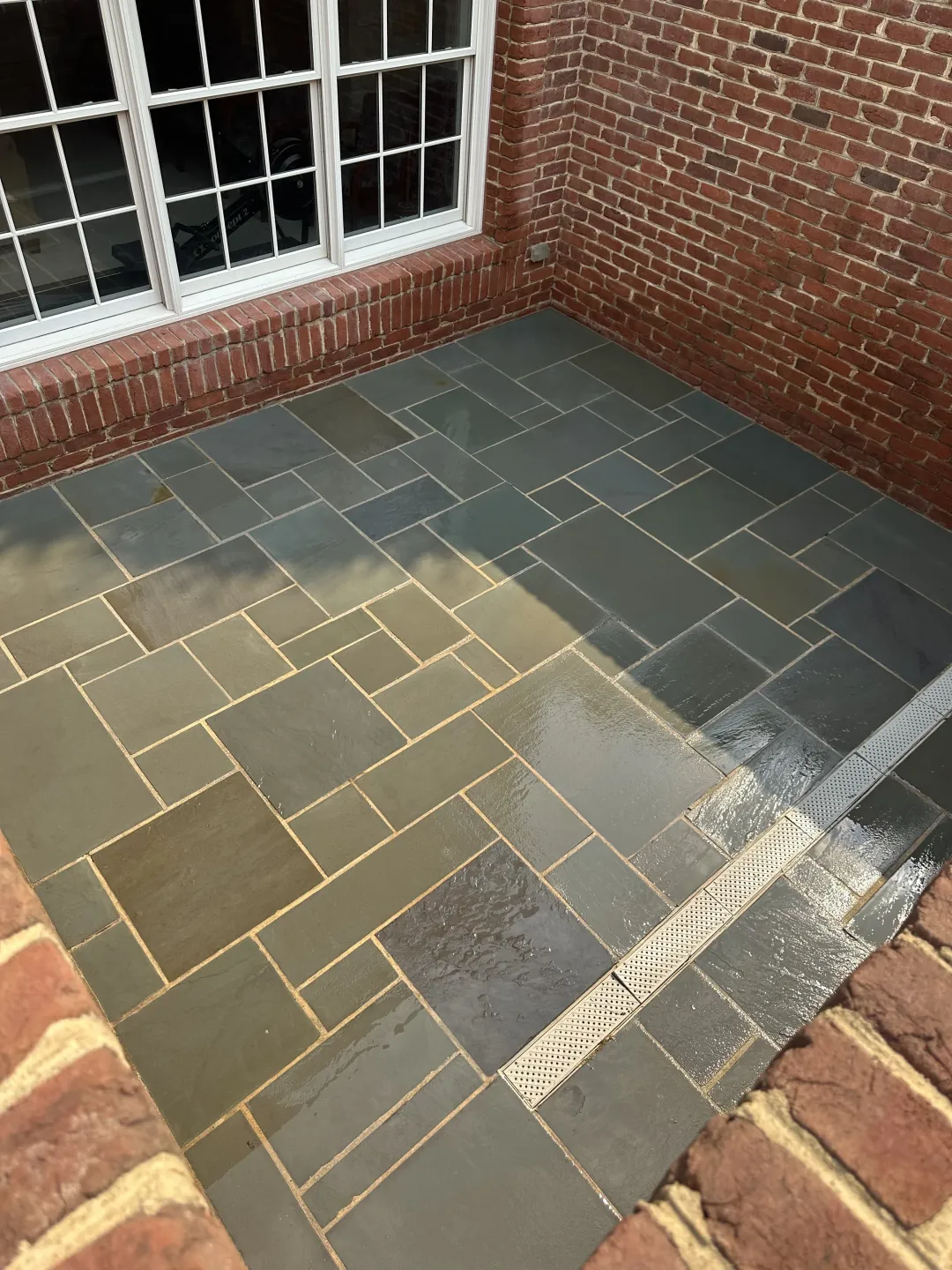 Brick patio with gray and tan pavers beside a brick wall and window, with a wet section and white residue on the floor