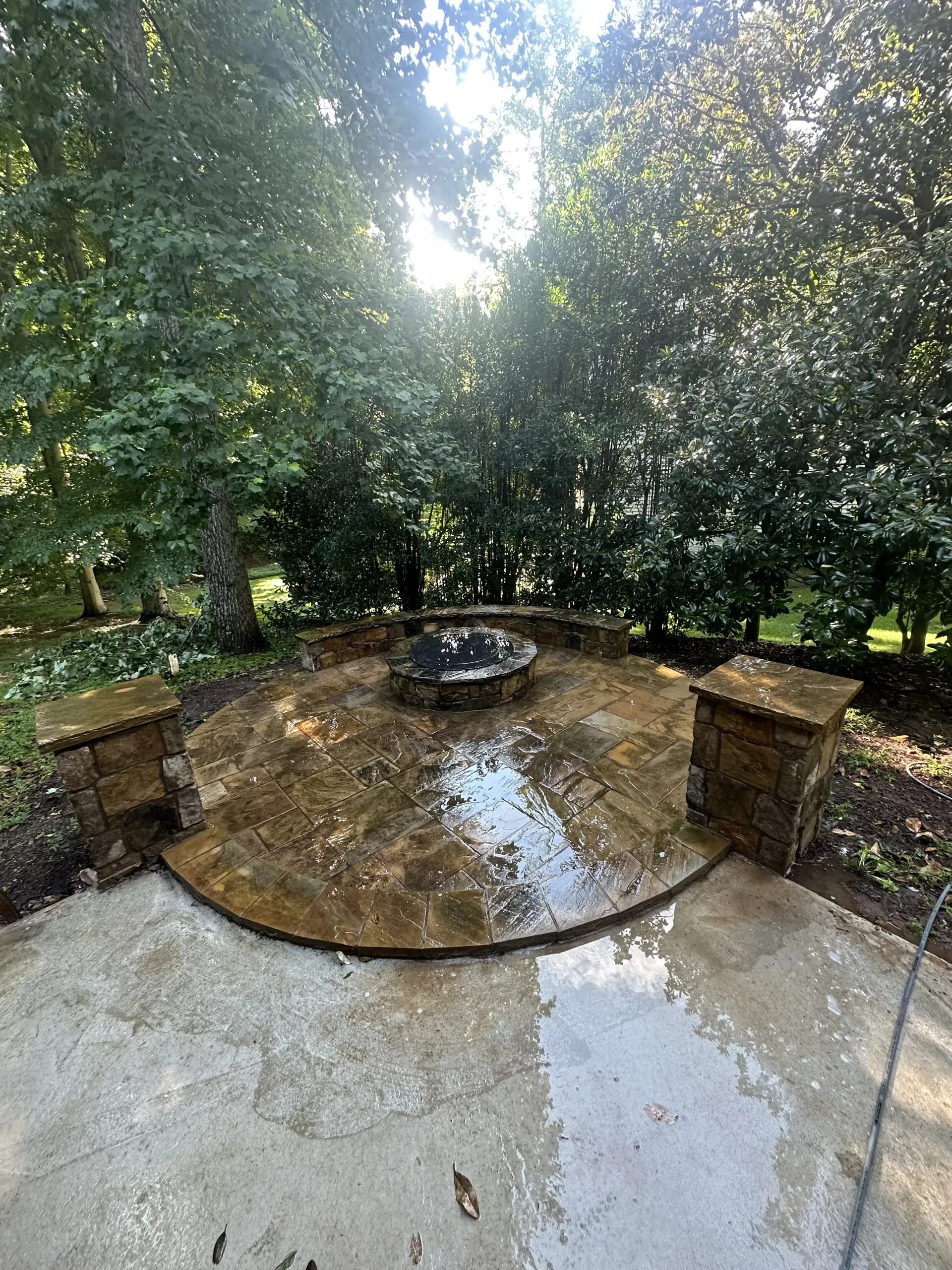 Outdoor stone patio with a circular fire pit, surrounded by lush trees and dense greenery.