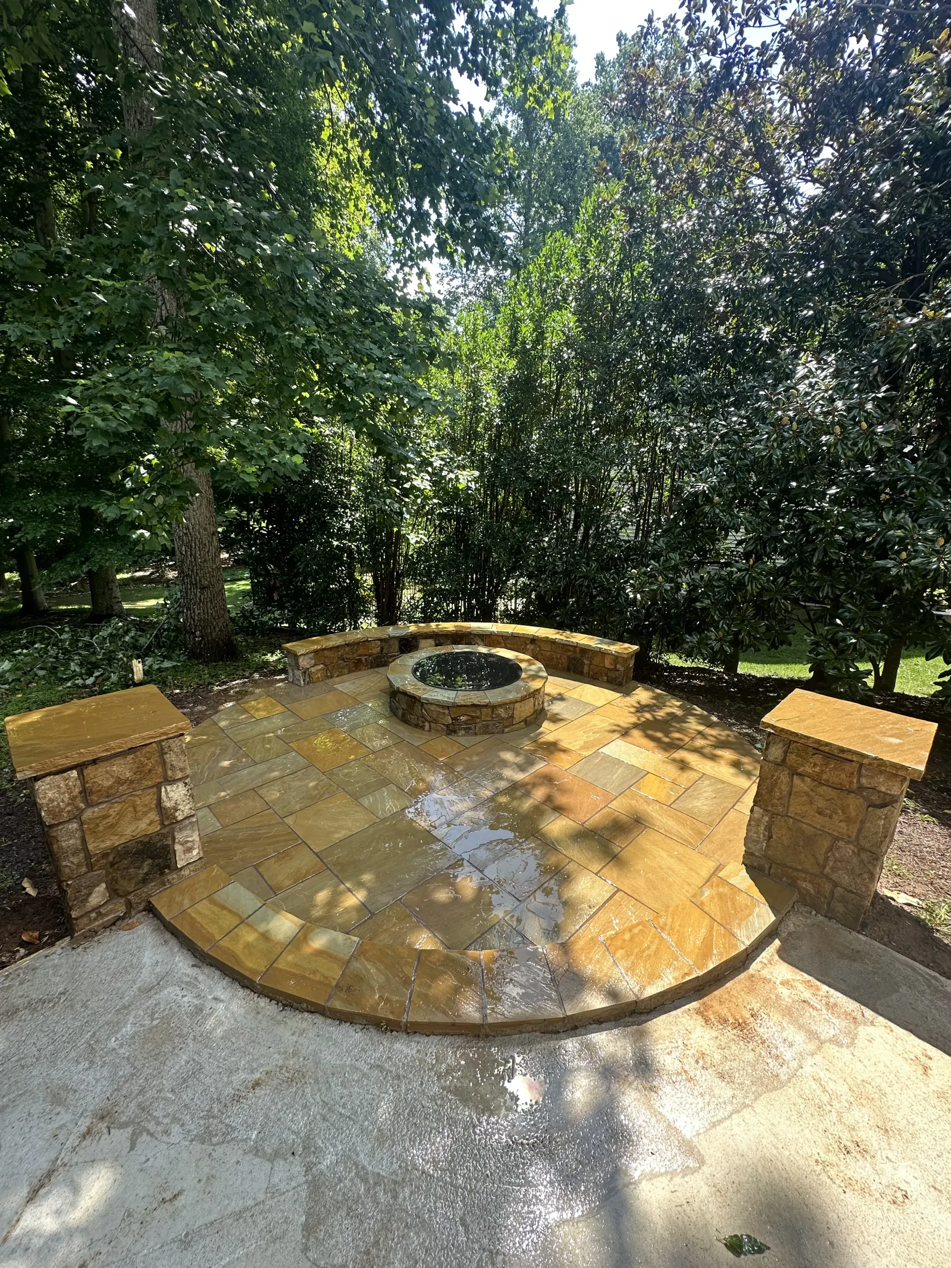Stone patio with a circular fire pit, bordered by brick pillars and surrounded by dense green trees.