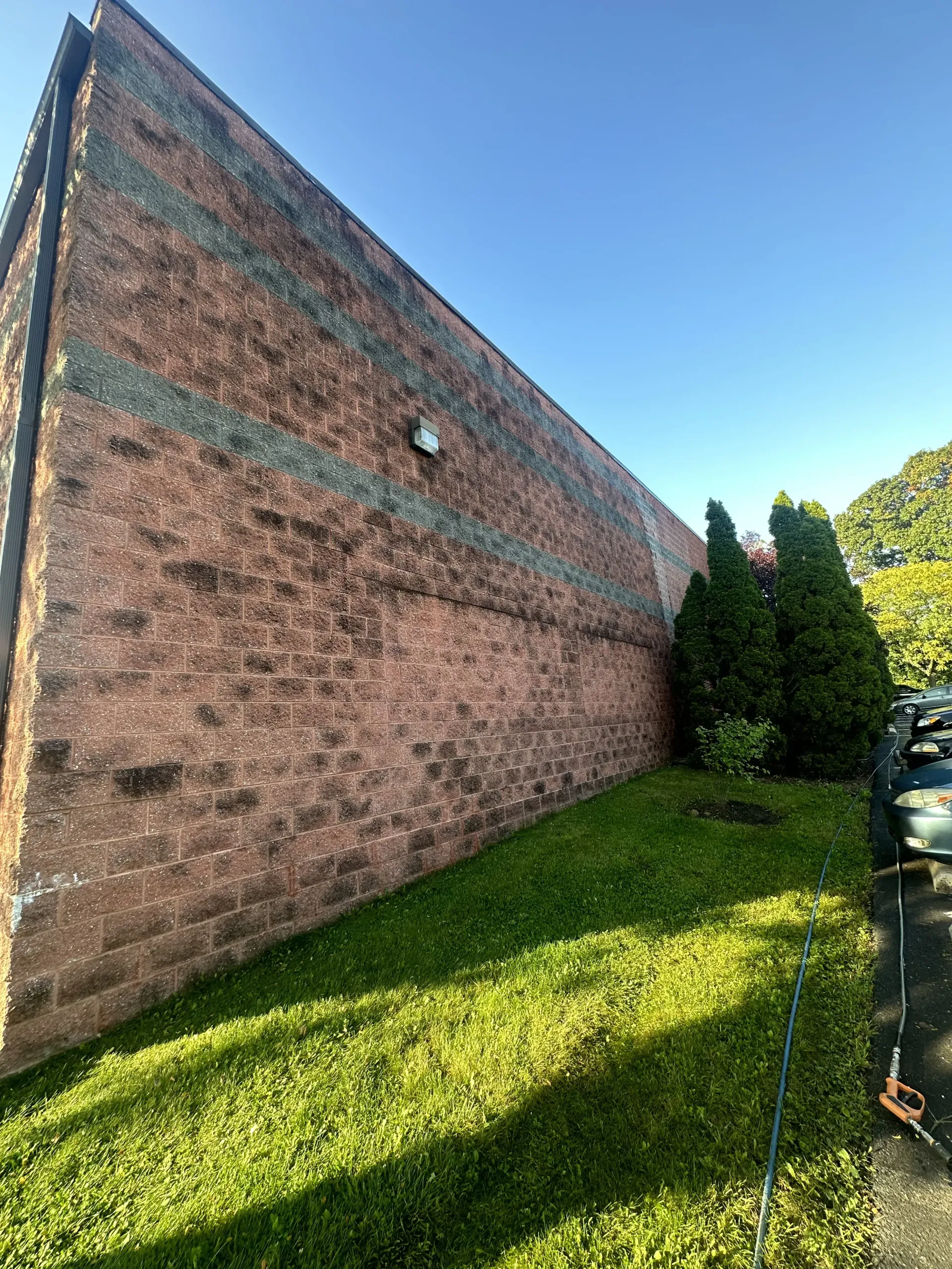 Long brick wall beside a grassy yard under a clear blue sky, with shrubs and a parked car at right.
