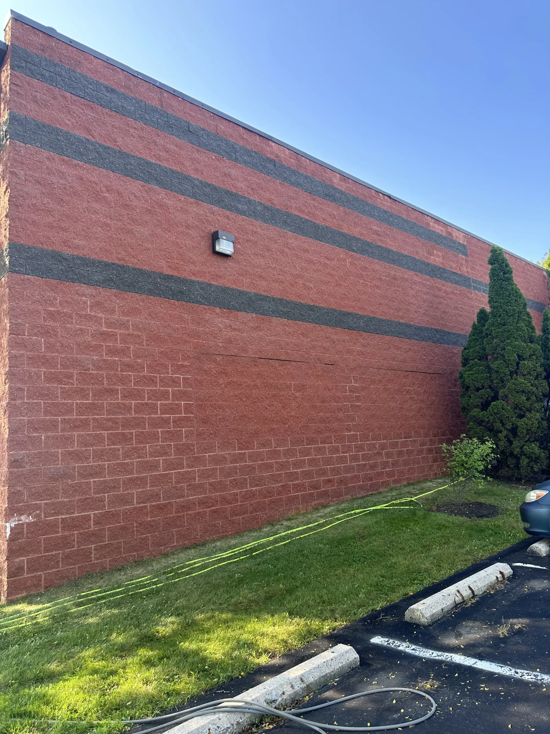 Red brick building wall with dark horizontal bands, a small light fixture, and grass beside a parking lot.