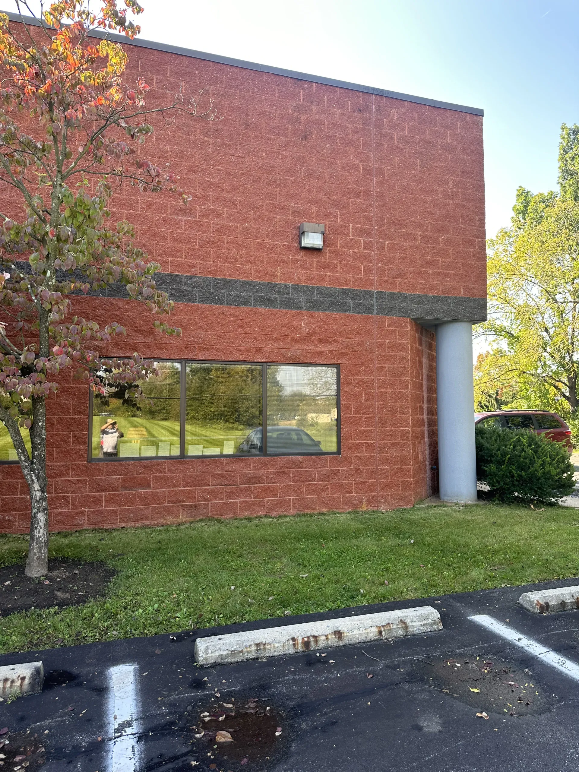 Red brick building exterior with a large window, white column, and trees beside a parking area.