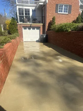 Driveway leading to a white garage beside brick houses and a black trash bin in sunlight