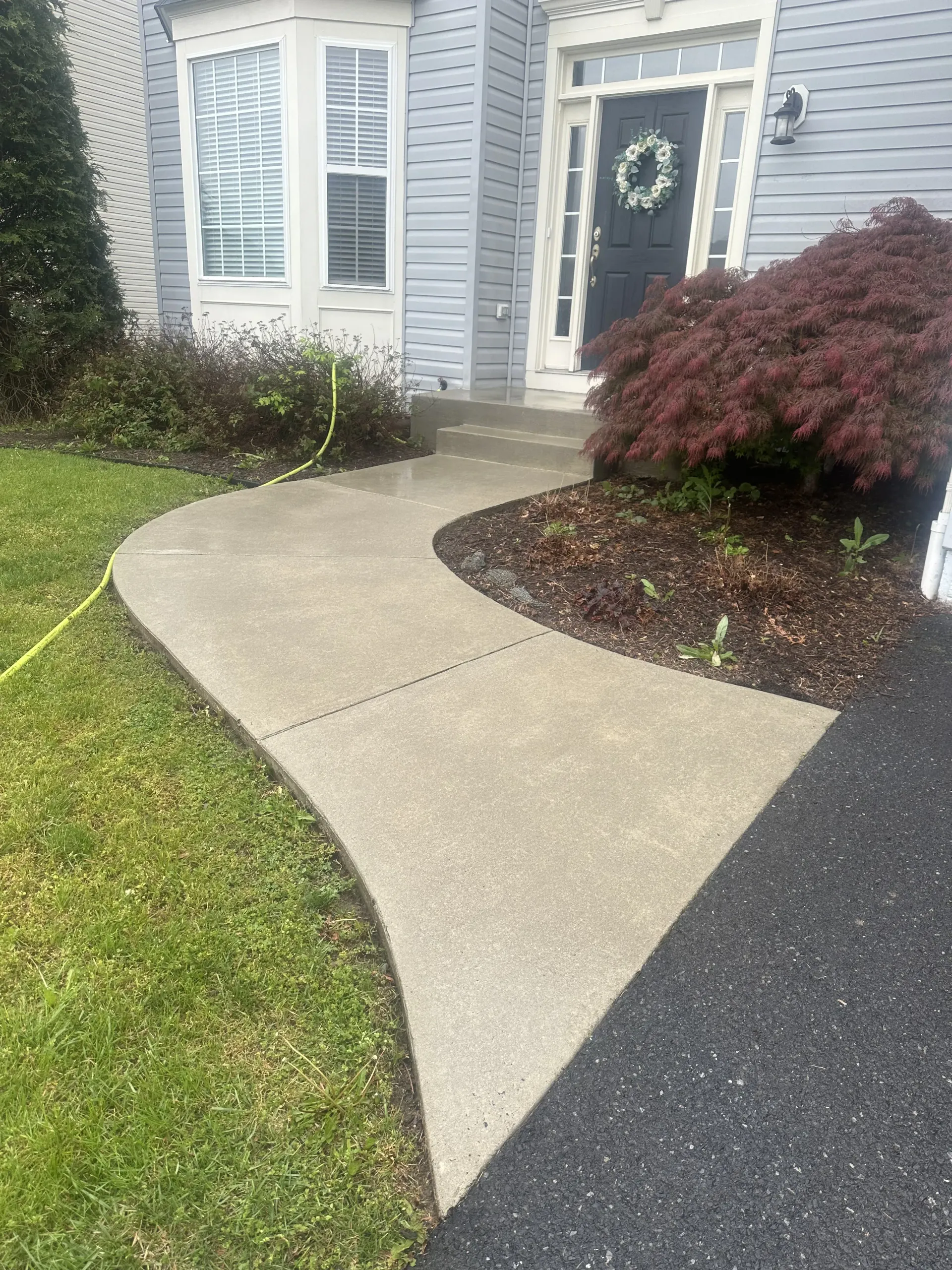 Curved concrete walkway leading to a front door with white-trimmed windows and shrubs beside a house