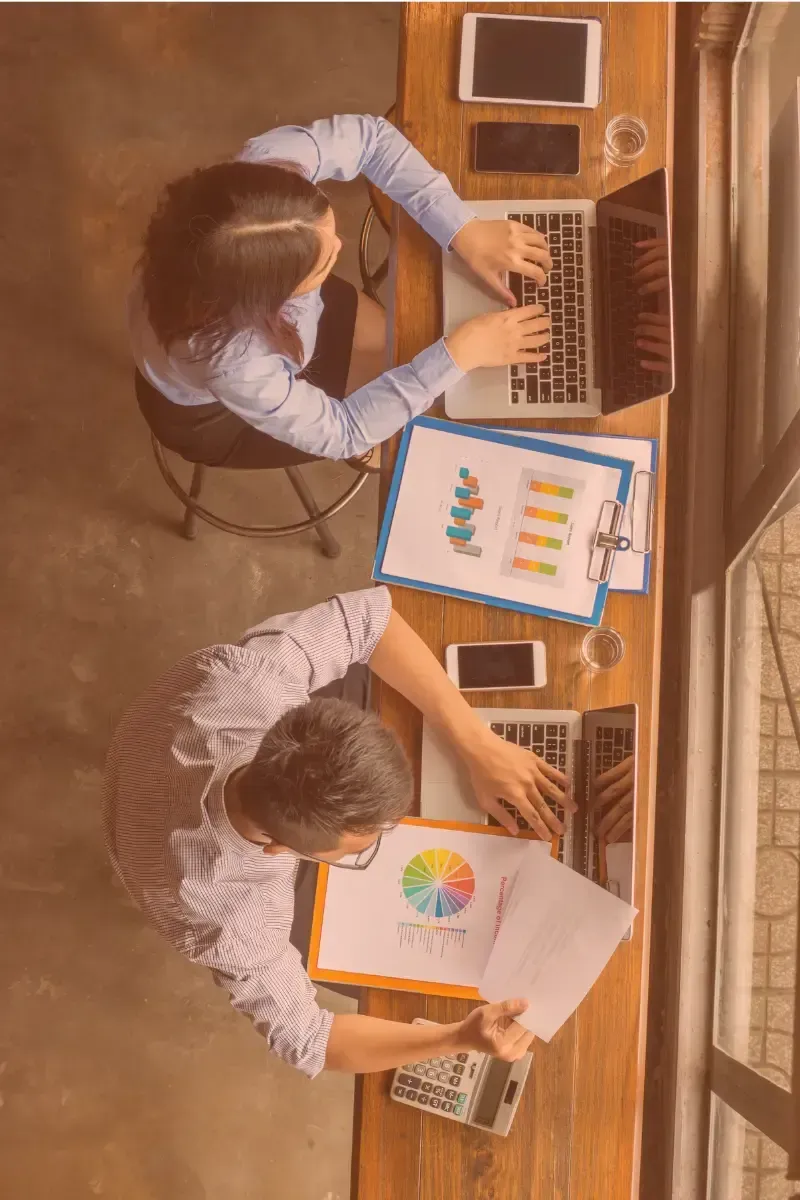 Two people working at a wooden table with laptops, charts, and mobile phones.