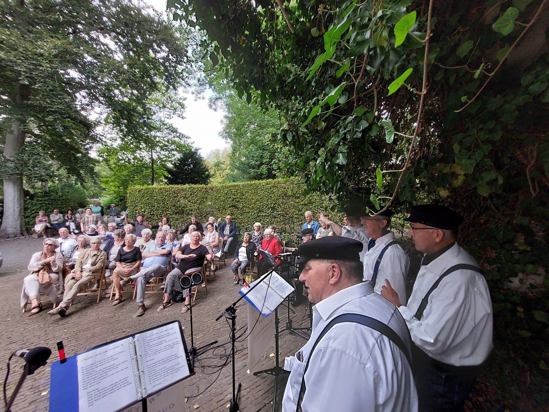 Two singers in white shirts and black berets perform for an outdoor audience seated in a park.