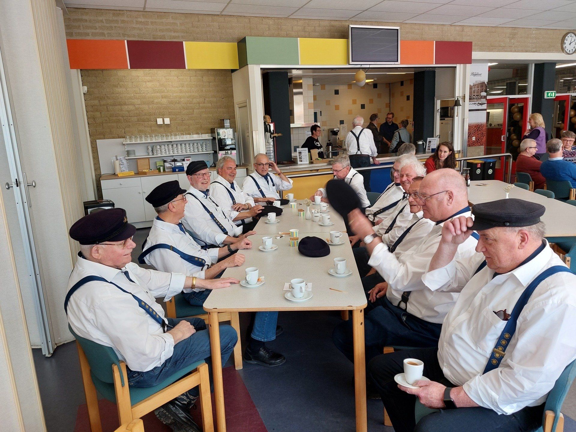 A group of people wearing hats and suspenders sit at a table in a brightly lit room, socializing over coffee.
