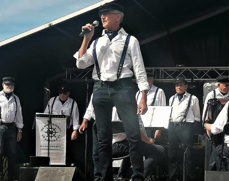 A performer sings into a microphone on a stage with a band, all wearing matching white shirts, dark suspenders, and caps.