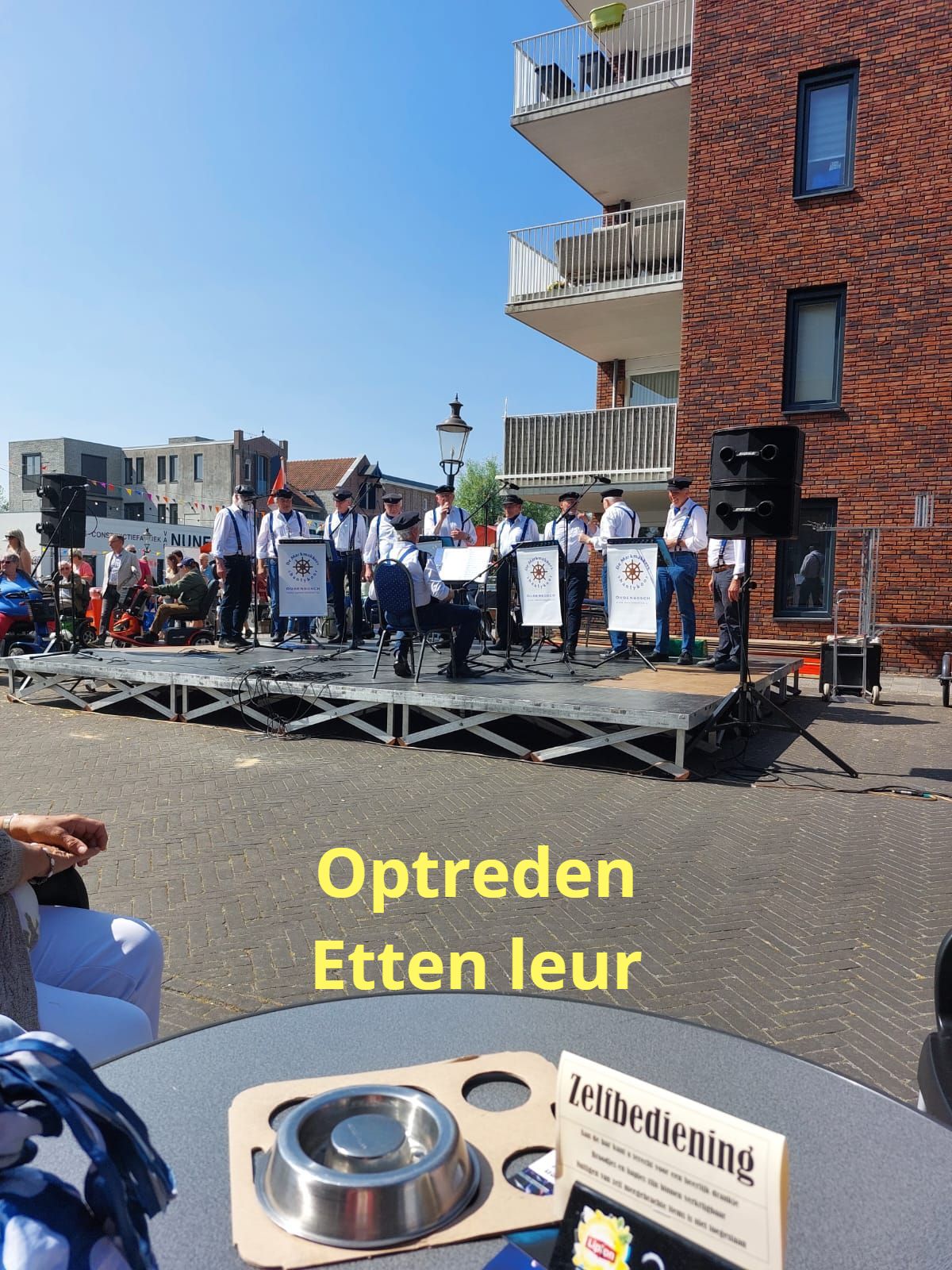 A band performs on a raised stage outdoors in Etten-Leur, viewed from a table with a self-service sign and metal bowl.