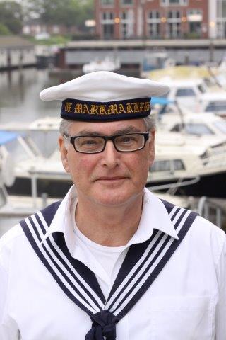 A person wearing a traditional sailor uniform and hat, standing in front of a marina with docked boats.